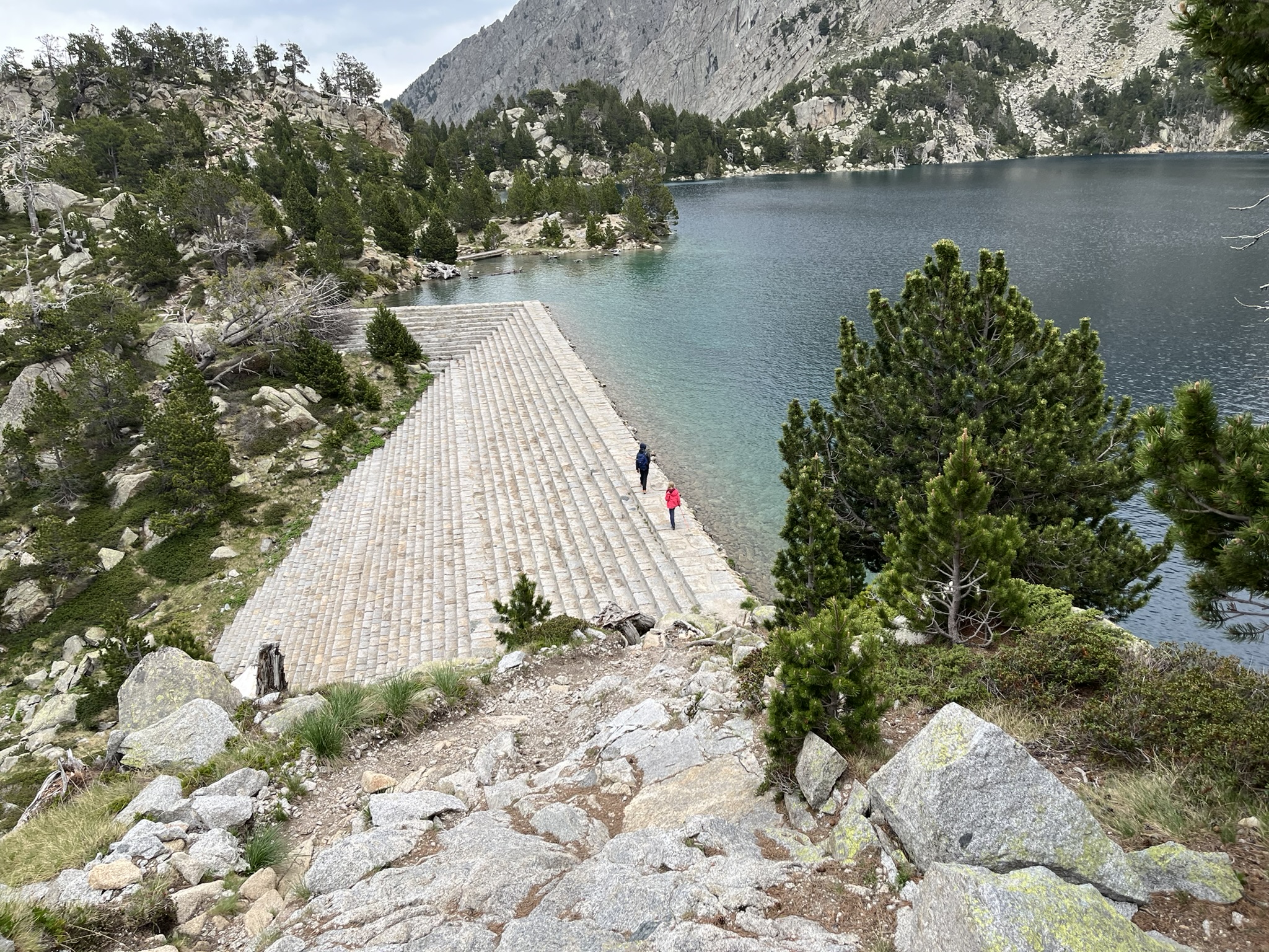 Two distant walkers on a stone embankment beside a turquoise mountain lake