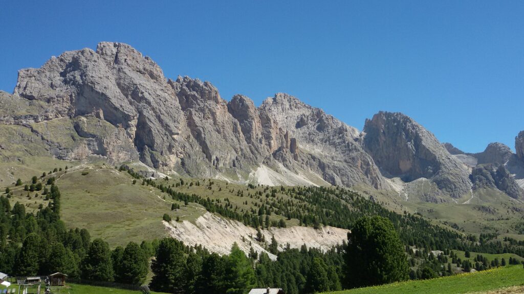 Pale Geisler rock towers above green meadows and pine trees seen from Col Raiser