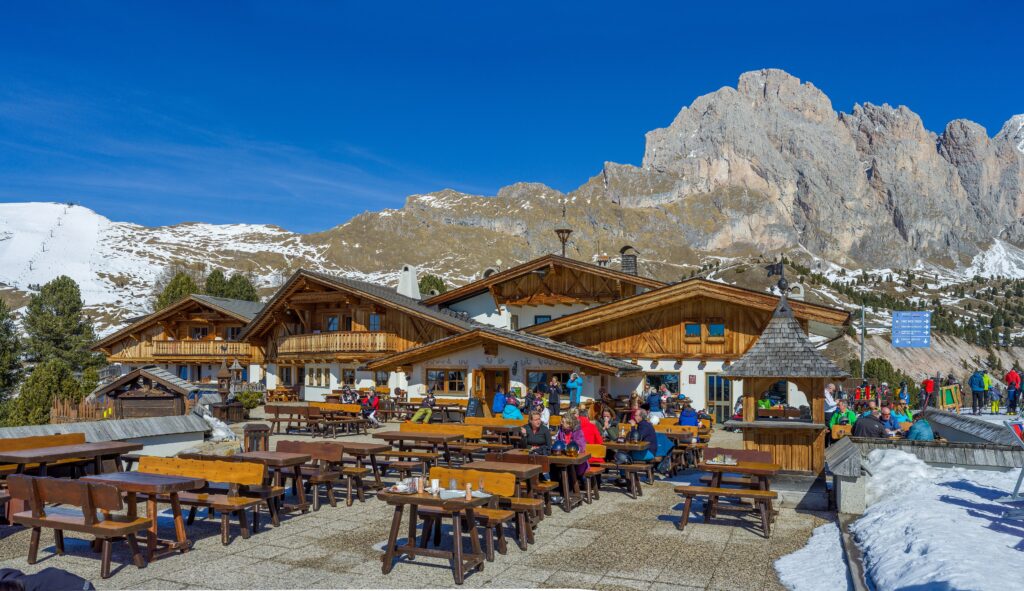 The timber balconies and terrace of Almhotel Col Raiser with snow patches and the Geisler peaks behind