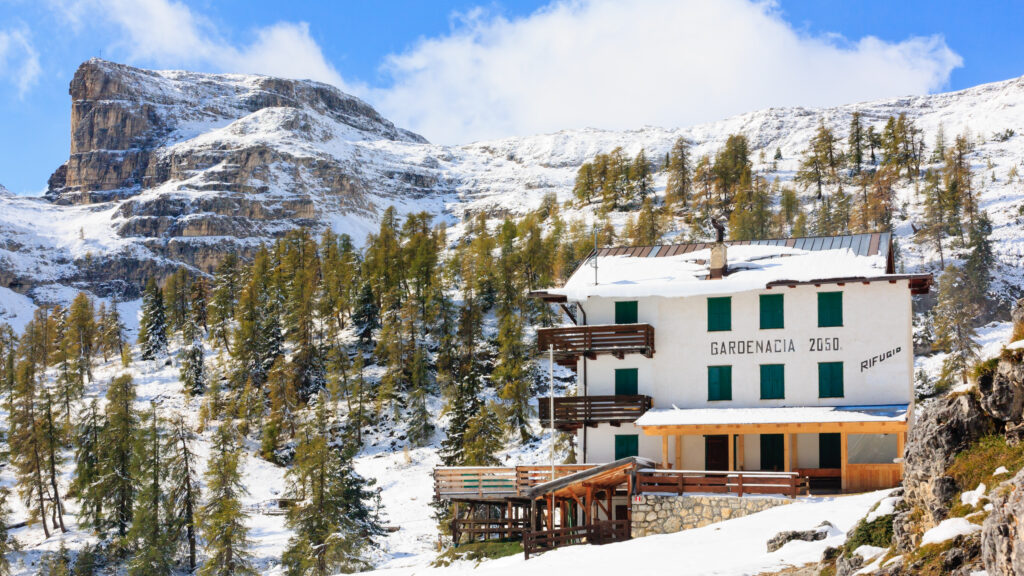White Rifugio Gardenacia with green shutters and wooden balconies below snow-covered limestone slopes and autumn larches